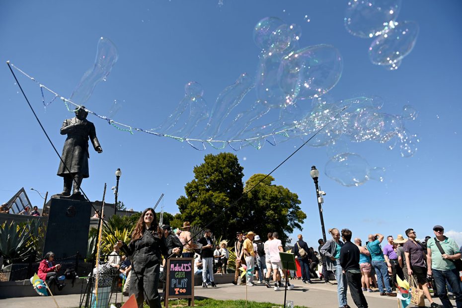 Woman creating large soap bubbles with a statue and crowd in the background on a sunny day.