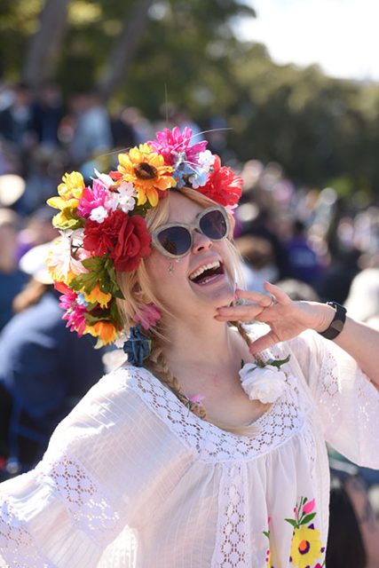 A woman wearing a floral headpiece and sunglasses laughs joyfully at an outdoor event.