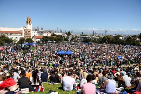 A large crowd gathered at an outdoor event on a sunny day with a city skyline in the background.