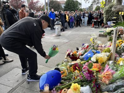 A person placing flowers at a makeshift memorial with onlookers in the background.