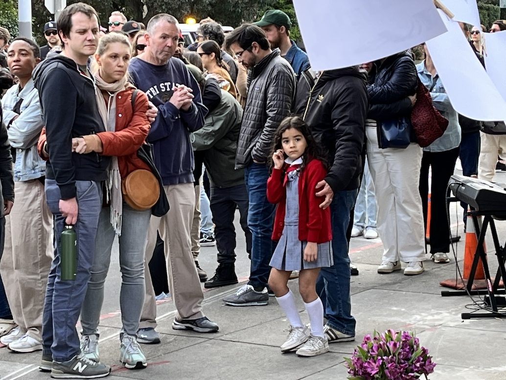 Crowd of people with various expressions attending an outdoor event, with a young girl in the foreground looking towards the camera.