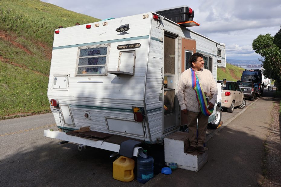 A person stands beside a parked travel trailer on a roadside, equipped with external water containers and a rear-mounted air conditioning unit, in a serene landscape with green hills in the background.