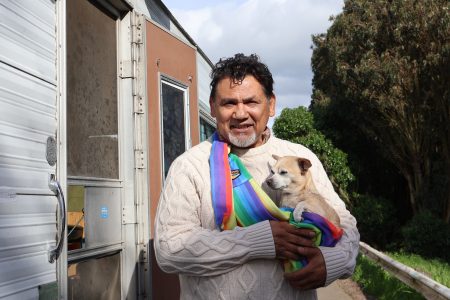 A man wearing a cream sweater and a colorful scarf stands outdoors holding a small dog wrapped in a matching scarf, with a trailer in the background.