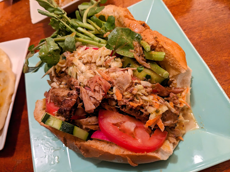 A plate with a pulled pork sandwich topped with slaw and tomato, accompanied by a side salad.