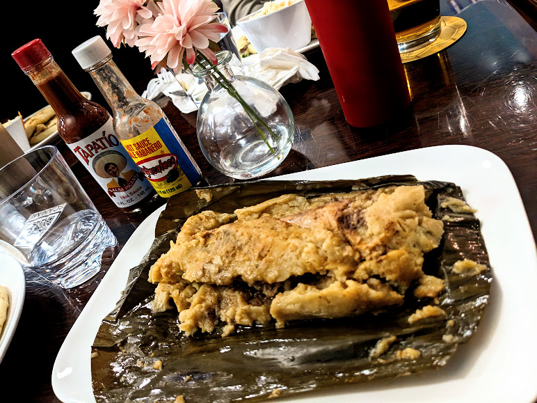 A traditional tamale unwrapped on a table surrounded by condiments and tableware.
