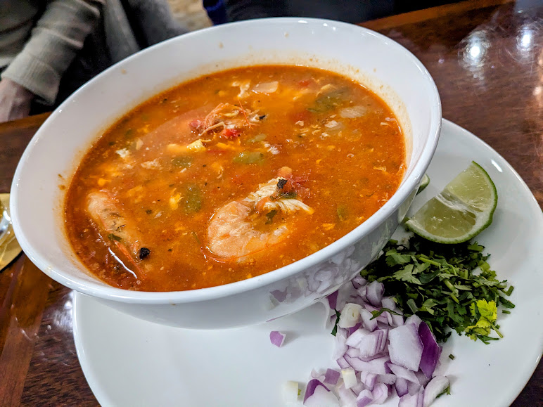 A bowl of spicy shrimp soup served with lime wedges and chopped onions and cilantro on the side.