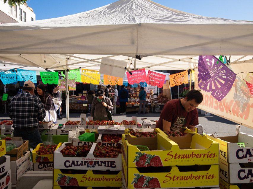 A bustling farmers market with colorful signs and fresh strawberries on display.