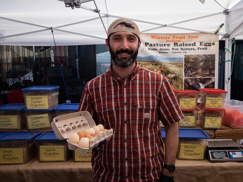 A smiling vendor at a farmer's market stall holding a carton of eggs with jars of nuts on display.