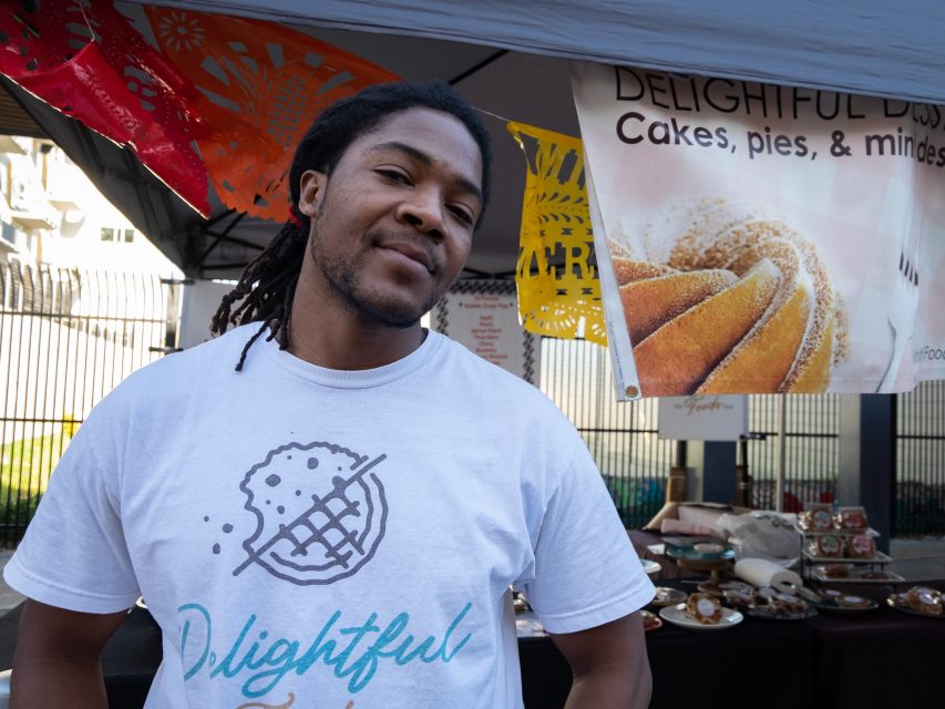 A smiling person wearing a t-shirt with the word "delightful" stands at a stall with cakes, pies, and signage in the background.