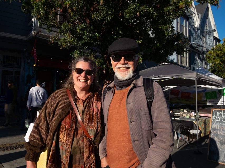 Two smiling people posing for a photo on a sunny street with market stalls and a building in the background.