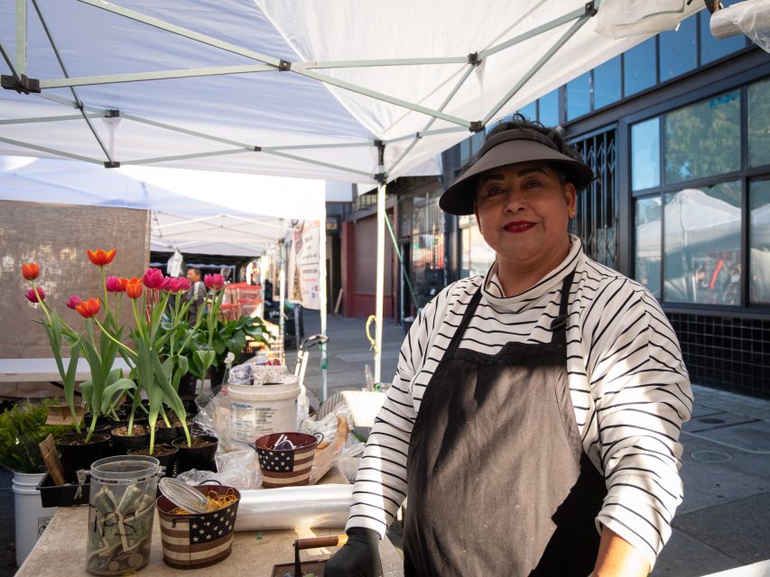 Vendor standing under a canopy at a market stall with flowers in the background.