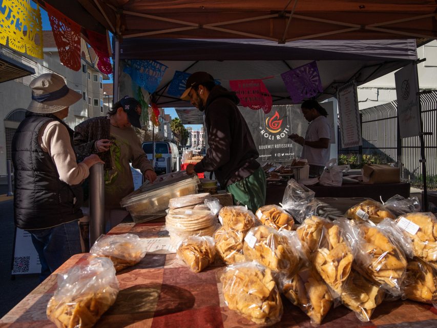People shopping at a street food stall with baked goods on display under a tent.