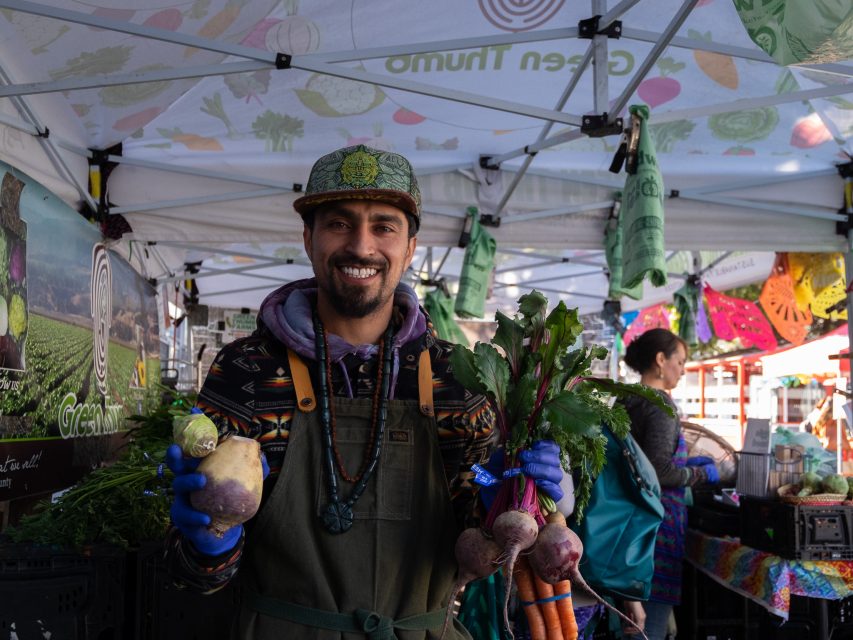 A man smiling at a farmer's market stall, holding a selection of fresh vegetables.