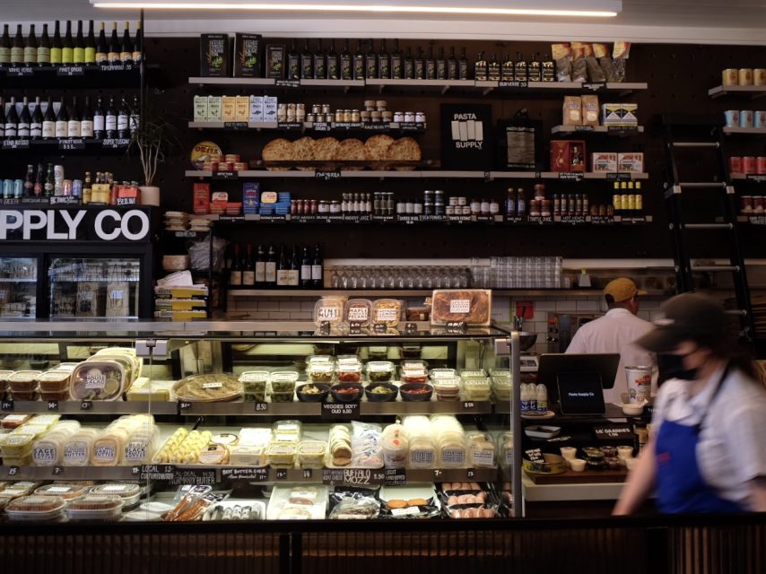 Pasta and deli counter with a variety of cheeses and packaged goods, attended by workers.