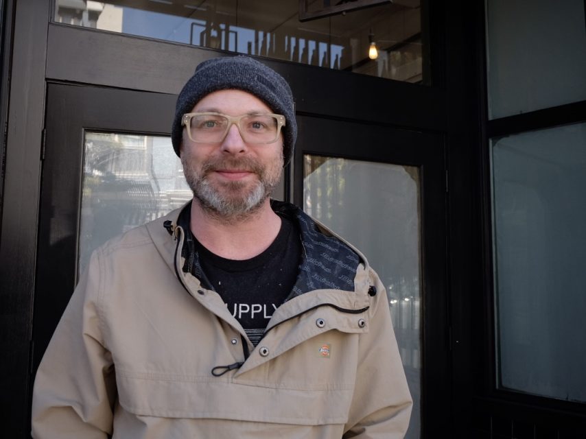 Man with glasses and a beanie smiling in front of a black door.