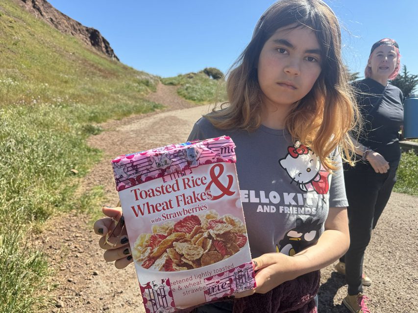 Young girl holding a bag of toasted rice and wheat flakes cereal with a woman in the background.