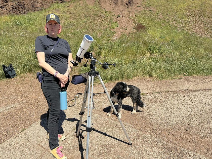 A person standing beside a telescope with a dog nearby on a sunny day with grassy terrain in the background.