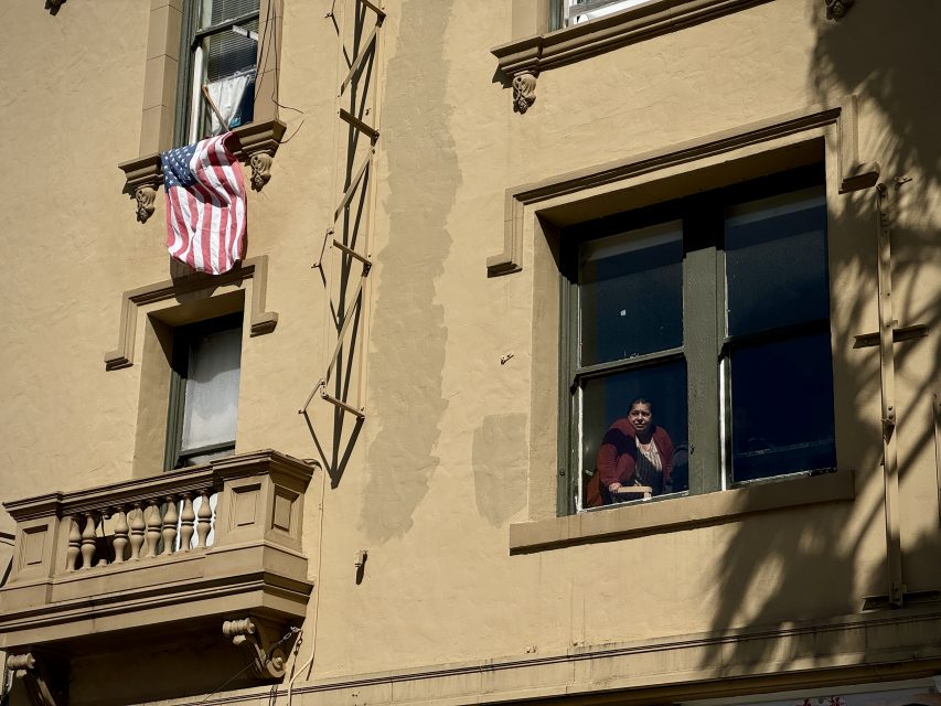 A person looks out from an apartment window beside a draped american flag.