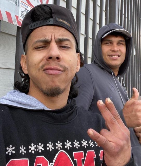 Two men posing for a selfie, one in the foreground making a Vargas Quero hand gesture and the other smiling in the background.