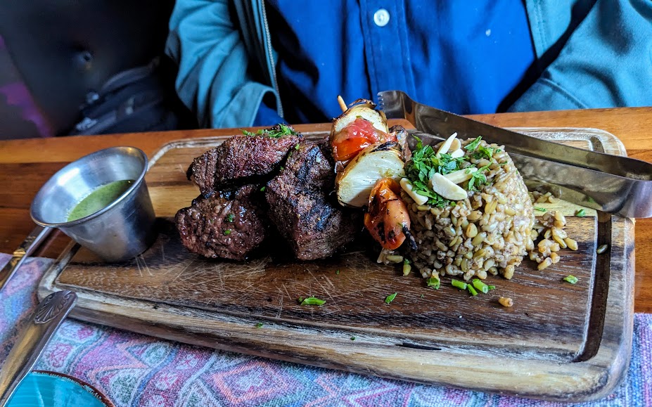 A person dining with a plate of grilled steak, rice, and vegetables served on a wooden board with a side of sauce.