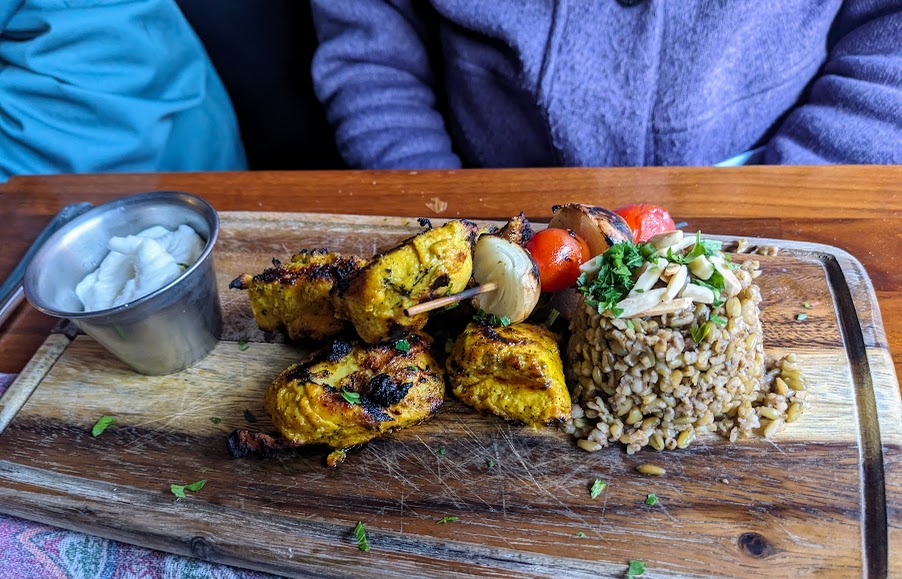 Grilled skewered chicken served with rice, vegetables, and a side of sauce on a wooden platter.
