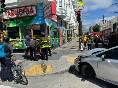 A silver sedan with damage to its hood in front of a taqueria with firefighters inspecting the damage from the crash