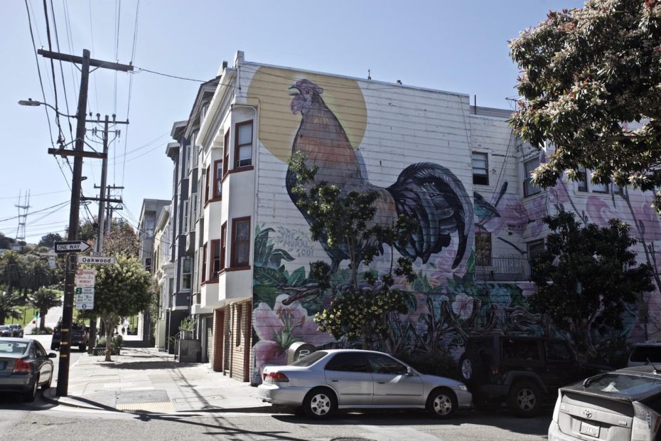 Urban street with a large mural of a rooster on a building's side wall.
