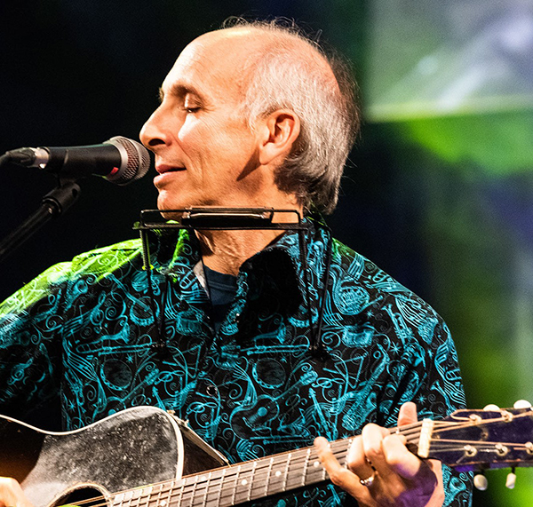 A balding man playing guitar and singing into a microphone with a harmonica mounted on a neck rack, wearing a patterned blue shirt.